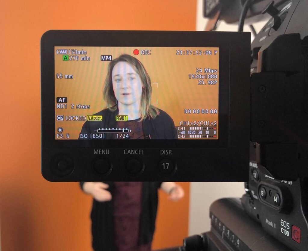 The blogger, Brenna, is standing in front of an orange wall and her face is mediated by the screen of a video camera.