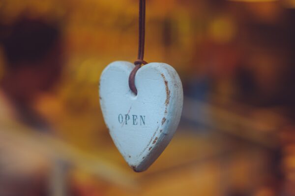 A white heart made of wood hangs from a string. The heart is printed with the word "open."