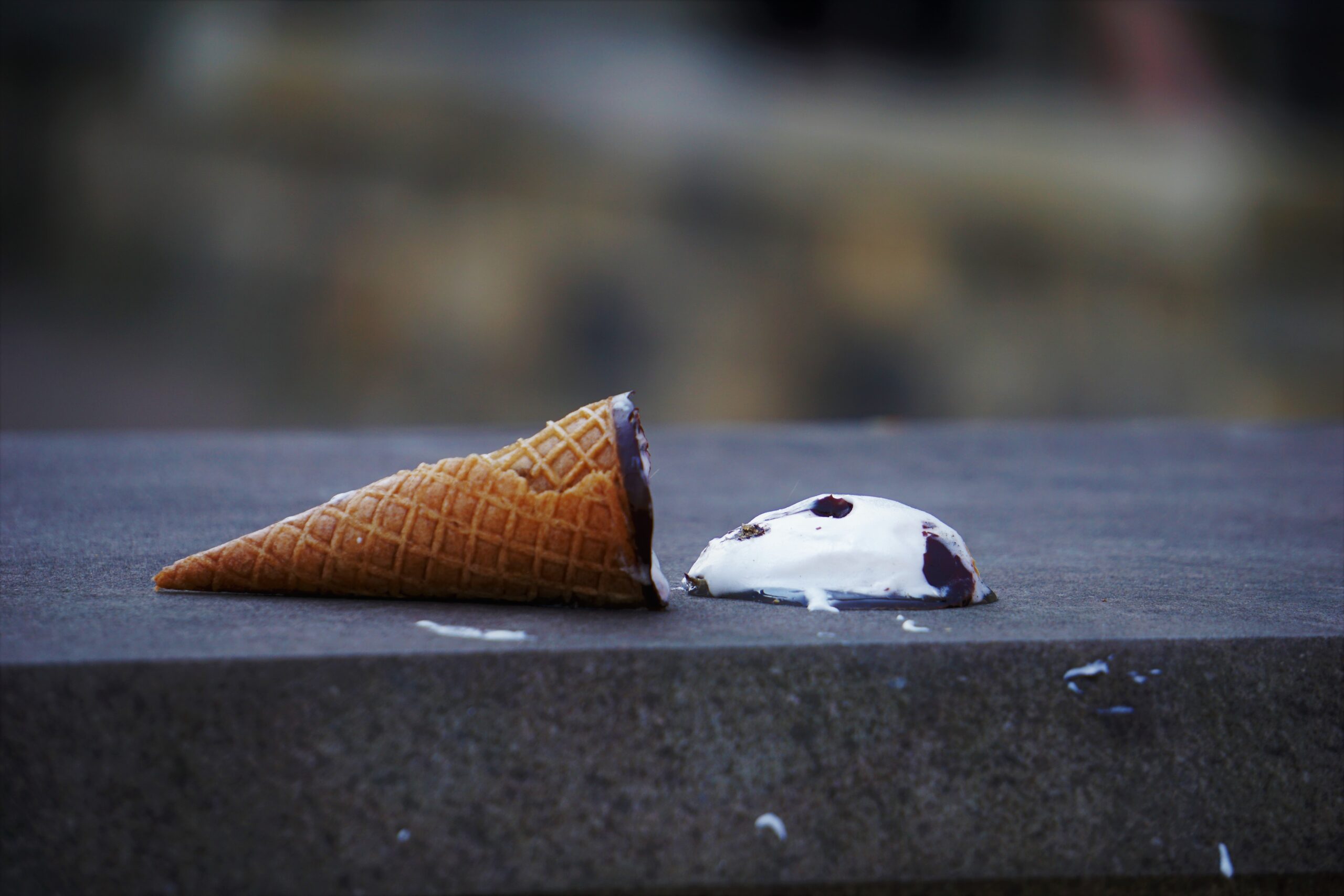 An ice cream cone lies on its side on the ground, the ice cream portion beginning to melt away.
