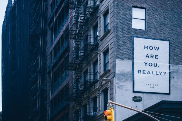 A sign on the side of a city apartment building reads, "How are you, really?"
